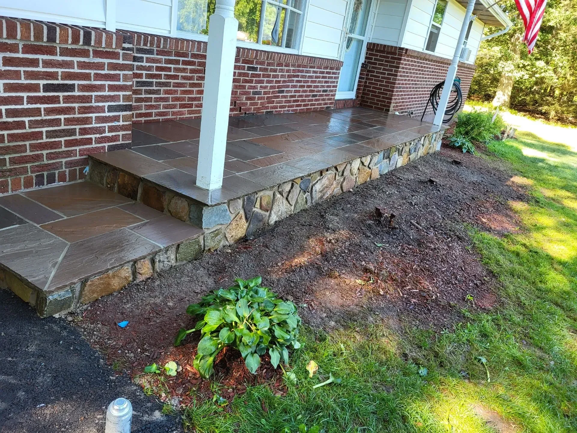 A newly constructed residential porch features a flagstone surface, matching steps, and a natural stone veneer base against a brick house exterior.