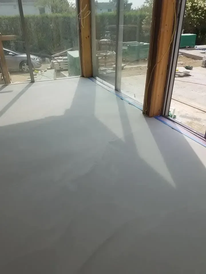 A newly poured, smooth gray floor is visible inside a room framed by large glass windows and wooden posts, with construction materials visible outside.