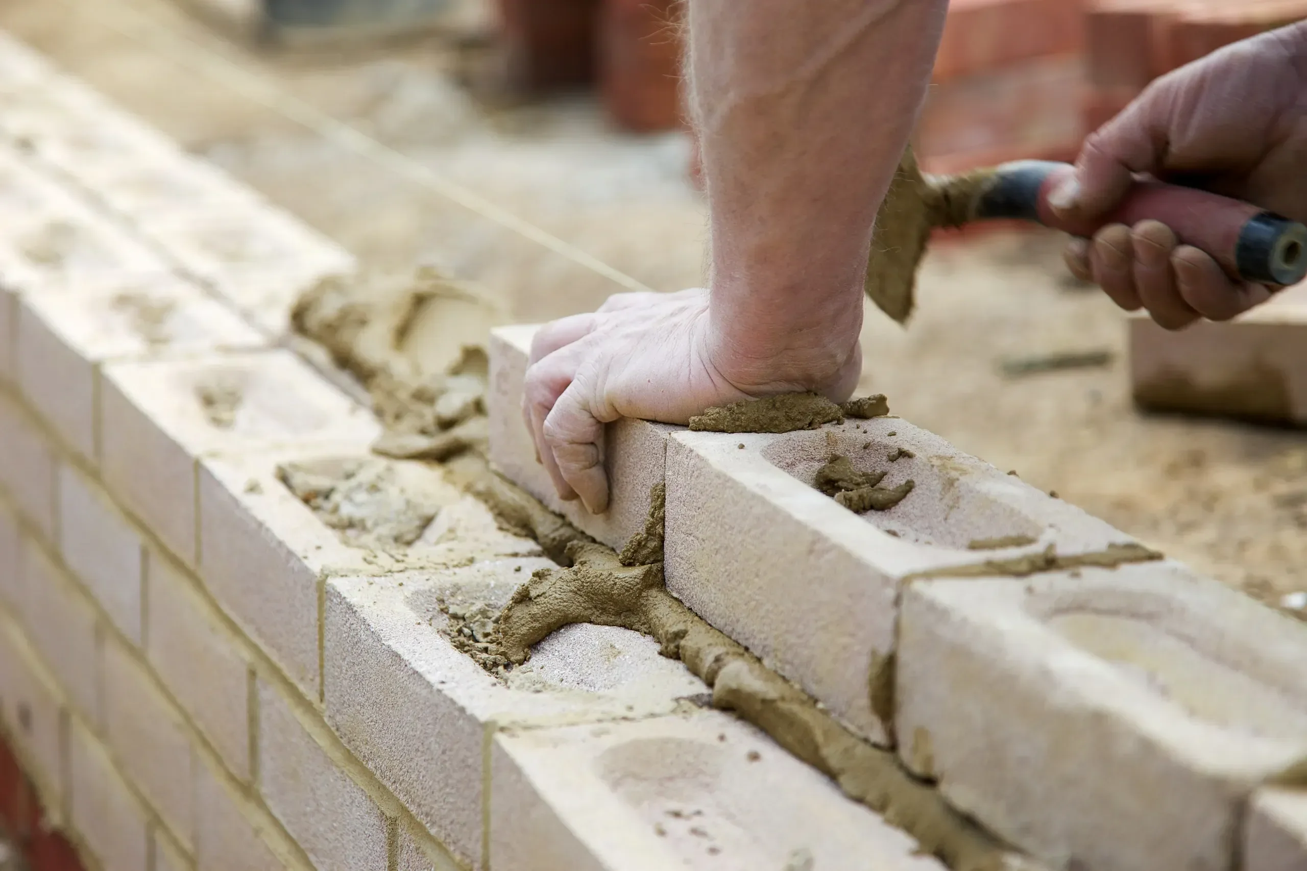 A construction worker uses a trowel to apply mortar while carefully setting a light-colored concrete block onto a partially built wall.
