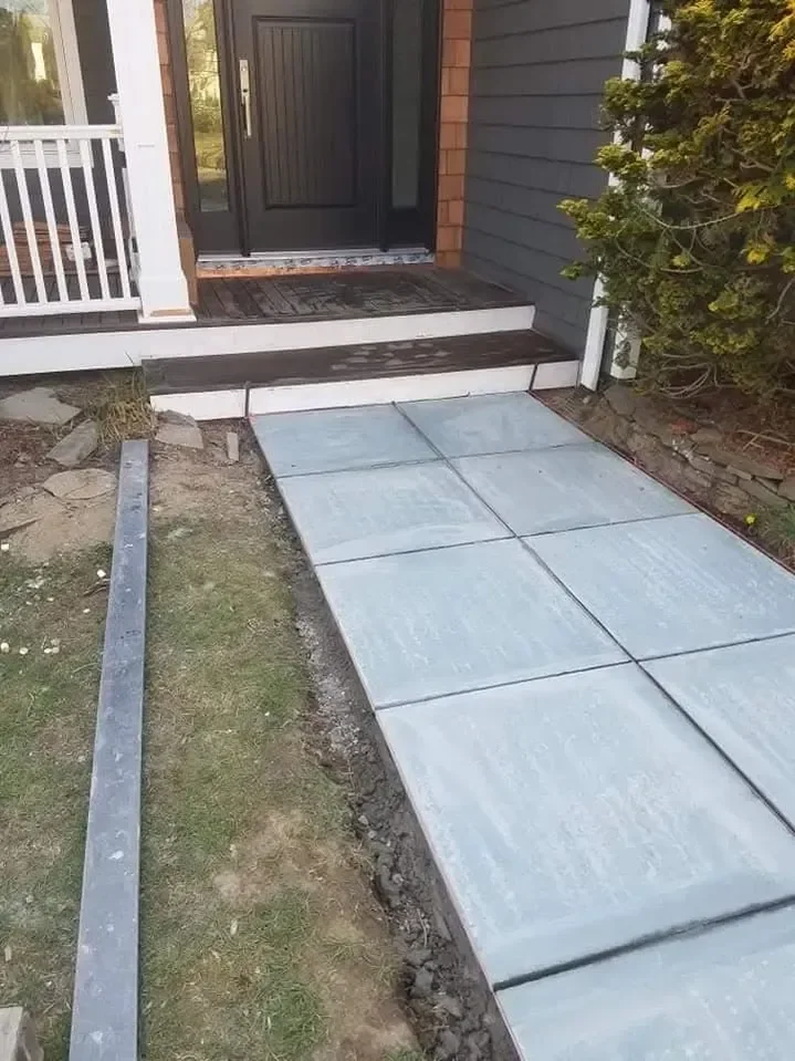 A newly installed segmented gray walkway leads up to the dark front door and wooden steps of a residential home, showing unfinished dirt edges.