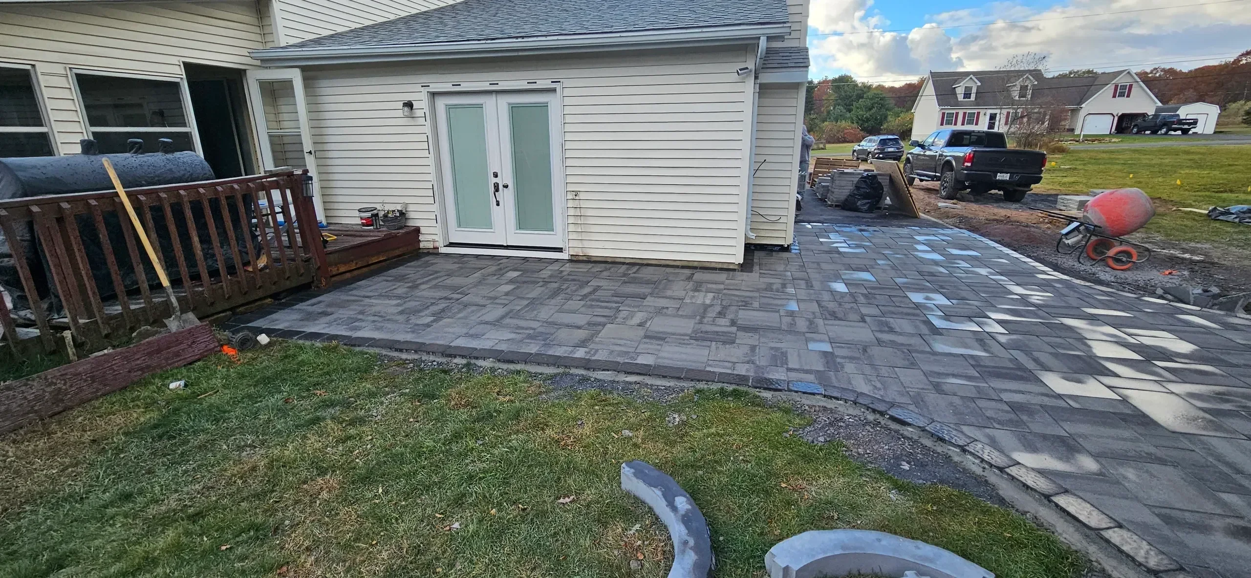 A newly installed gray paver patio and walkway extends from the back of a beige house, with construction materials and tools visible nearby.