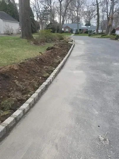 Newly installed stone edging separates a paved surface from a mound of disturbed soil and grass in a suburban setting.