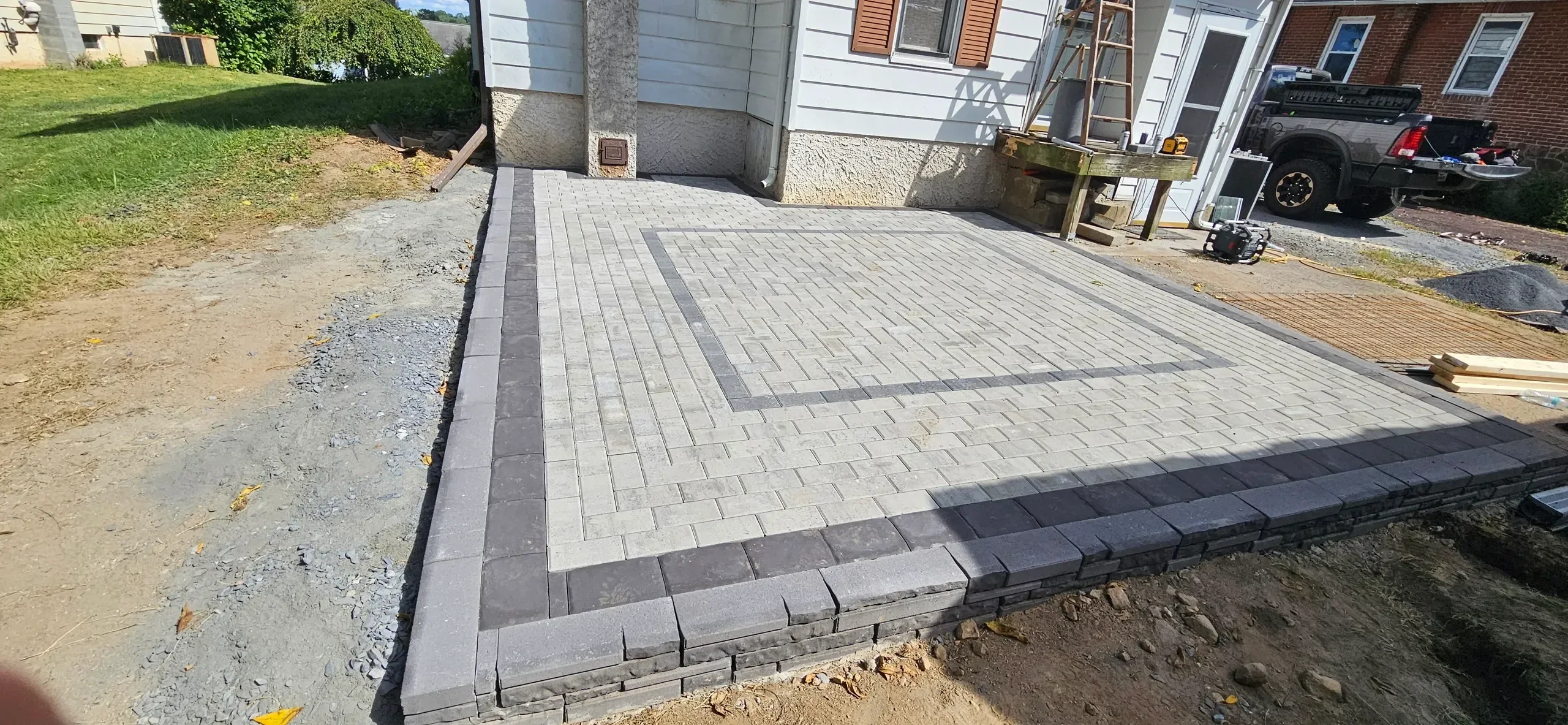 A newly constructed residential paver patio featuring light gray stones and a dark gray border design is shown adjacent to a house foundation amidst ongoing construction work.