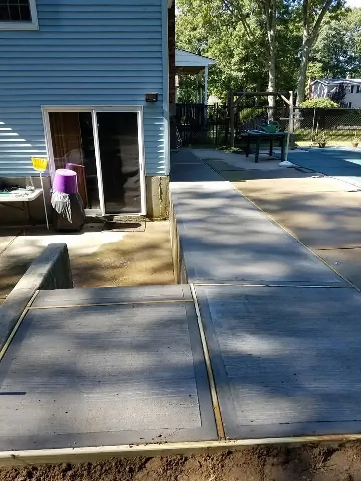 A newly installed textured concrete walkway leads away from a blue-sided house toward a fenced backyard area under bright sunlight.