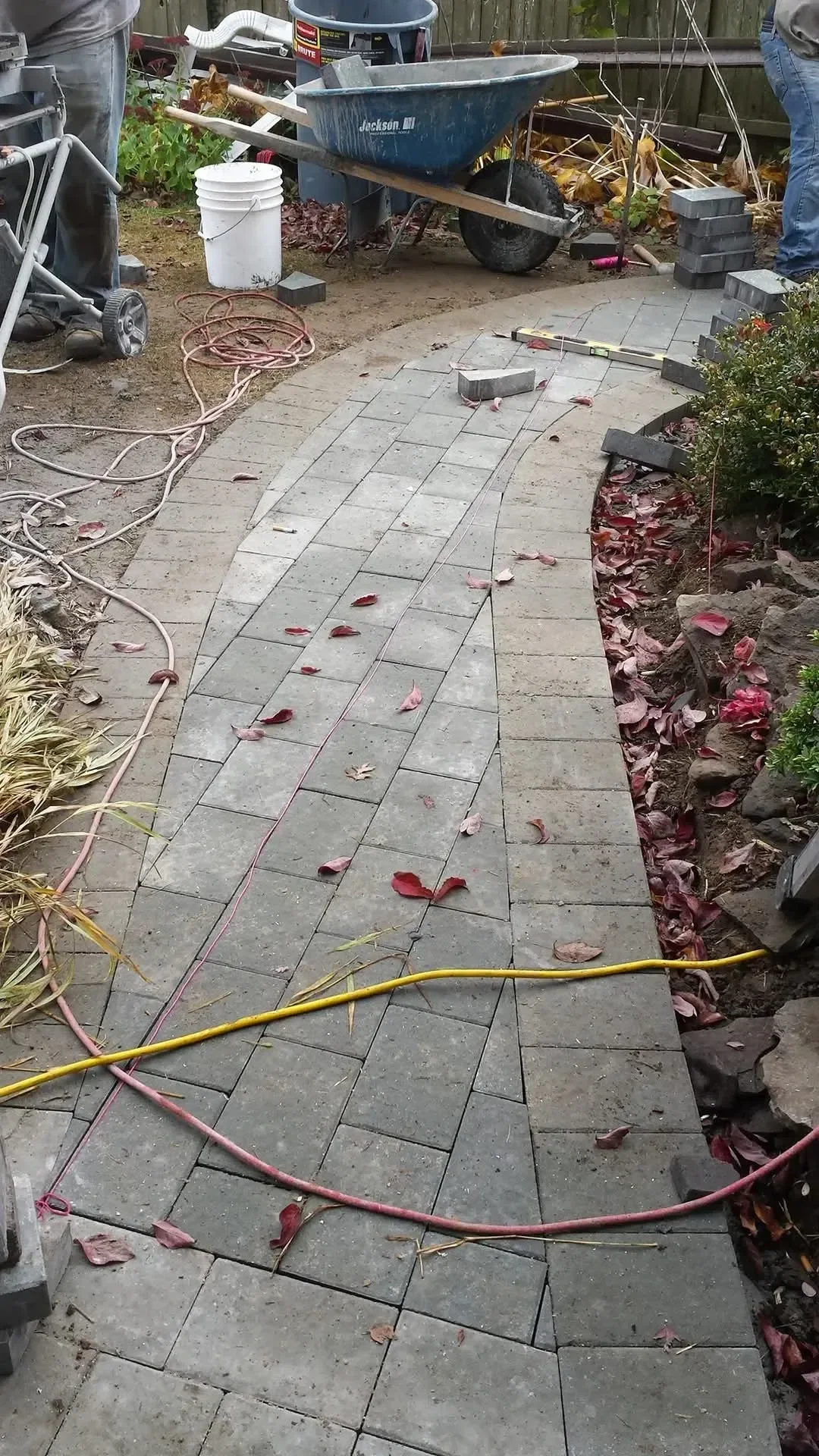 Construction workers are installing a curved gray paver walkway, surrounded by tools, materials, and autumn leaves on the ground.