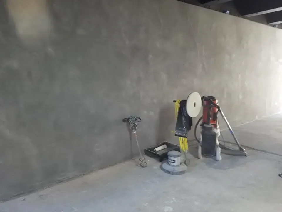 Various floor polishing and finishing tools, including a large red machine and yellow cables, rest against a long, textured gray wall in an interior space.