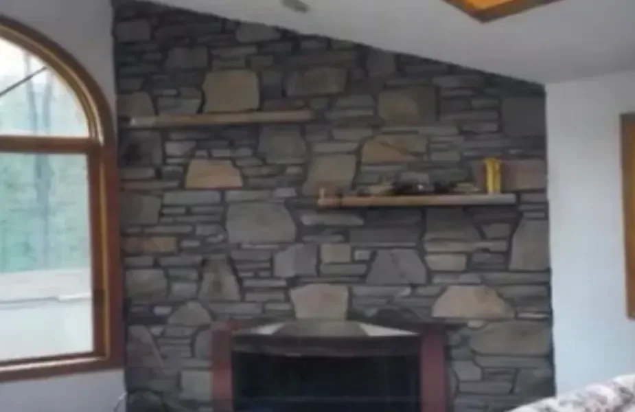 This completed interior features a rustic fireplace wall clad in irregular natural stone, accented by two wooden shelves and a large arched window overlooking trees.