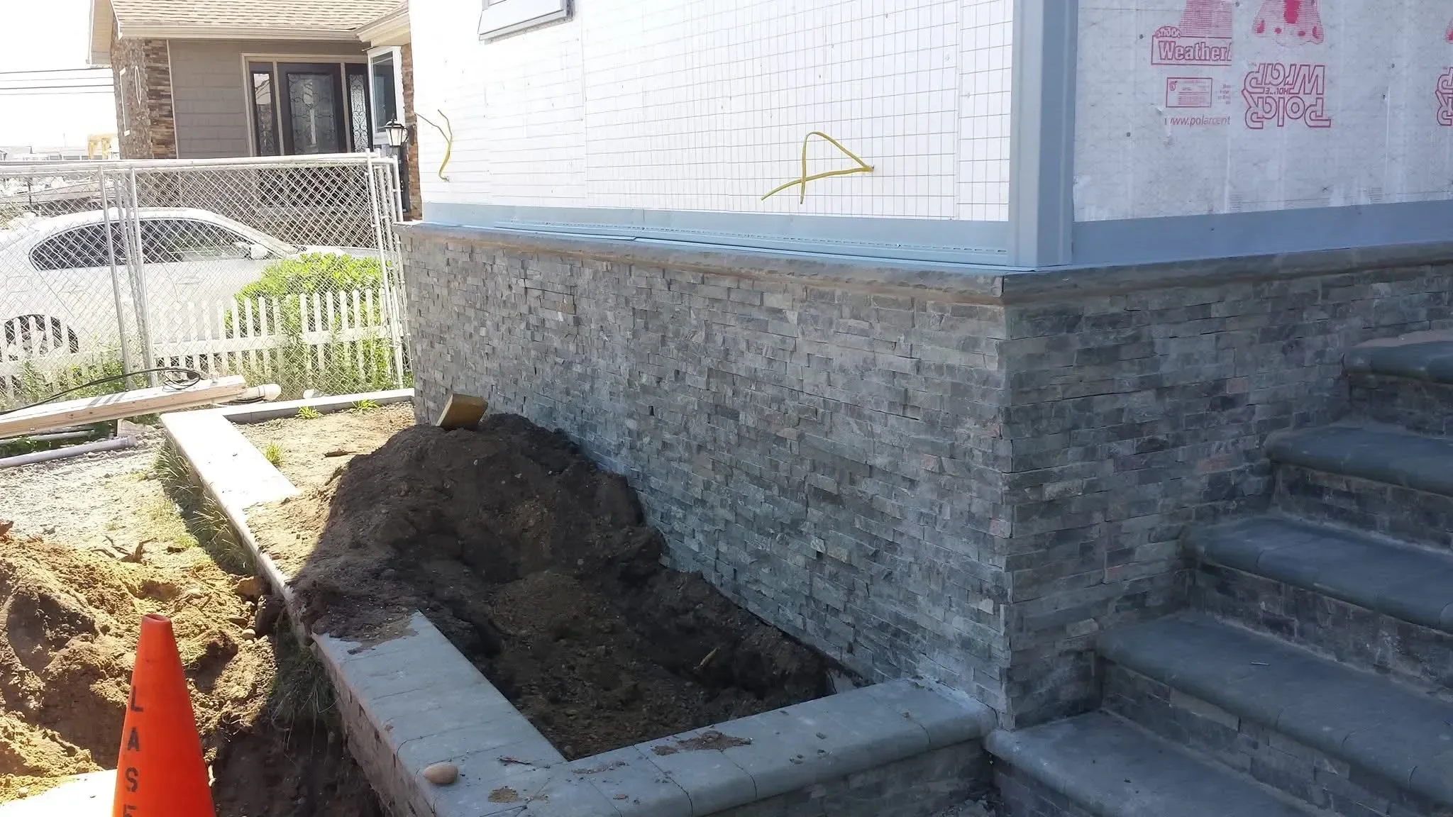 A residential foundation is being updated with dark gray stacked stone veneer cladding, while the upper walls remain unfinished, surrounded by excavated dirt and construction materials.