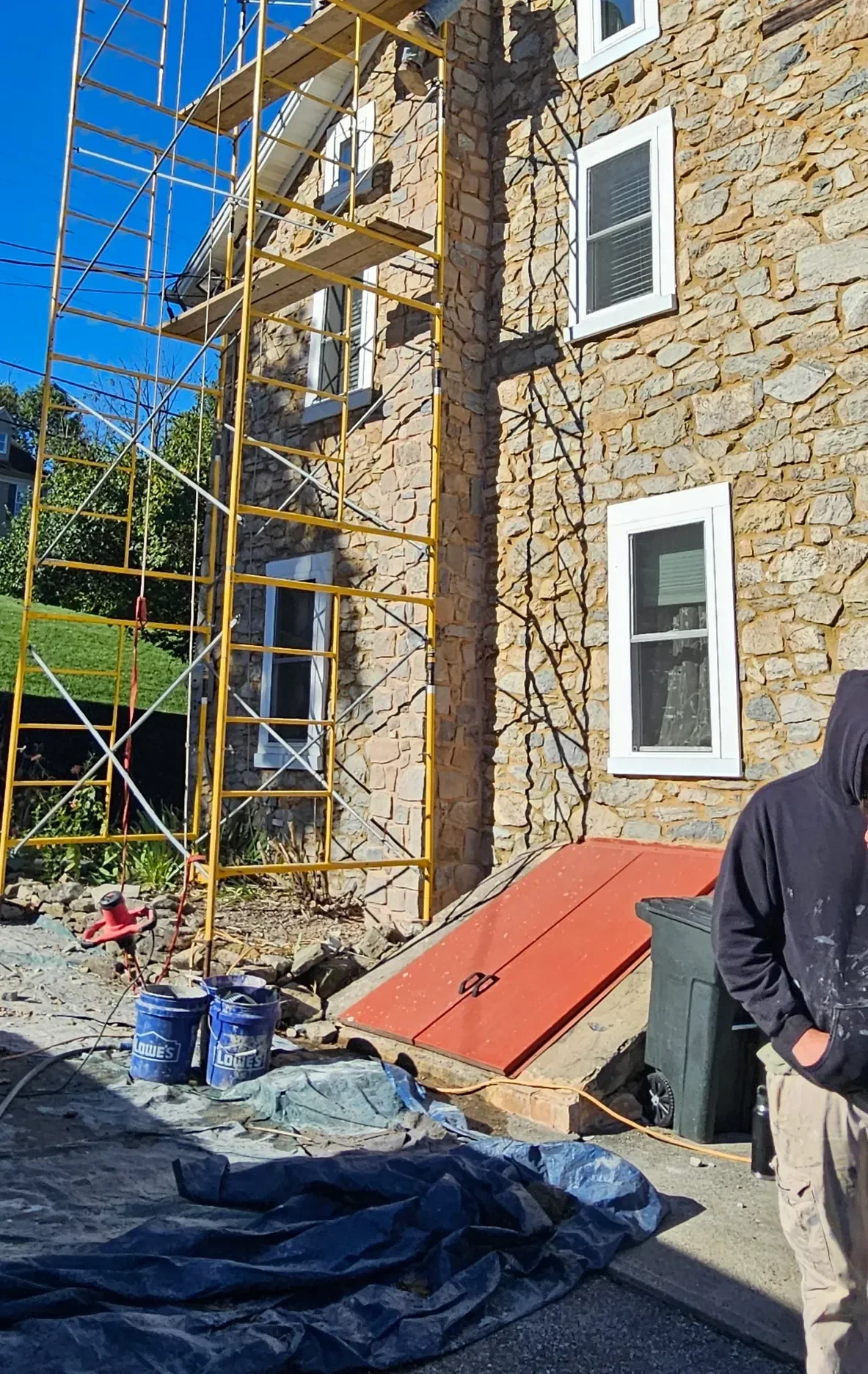 Yellow scaffolding stands against the stone exterior of a building undergoing renovation, with a worker and construction tools visible on the ground below.