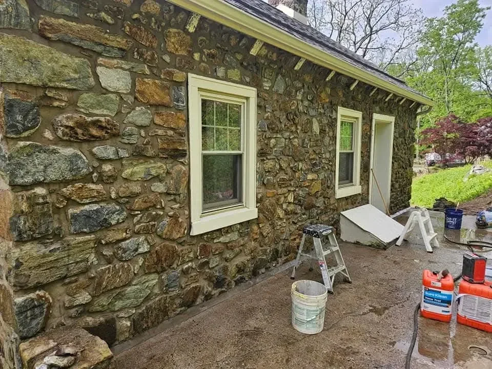 Restoration work is underway on the exterior of a building featuring rustic fieldstone masonry walls, visible by the presence of cleaning supplies and a ladder on the wet concrete patio.
