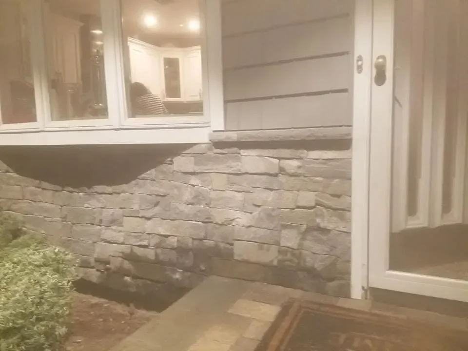 A nighttime view captures the exterior of a home featuring gray stacked stone veneer, horizontal siding, and a large window reflecting a brightly lit interior kitchen.