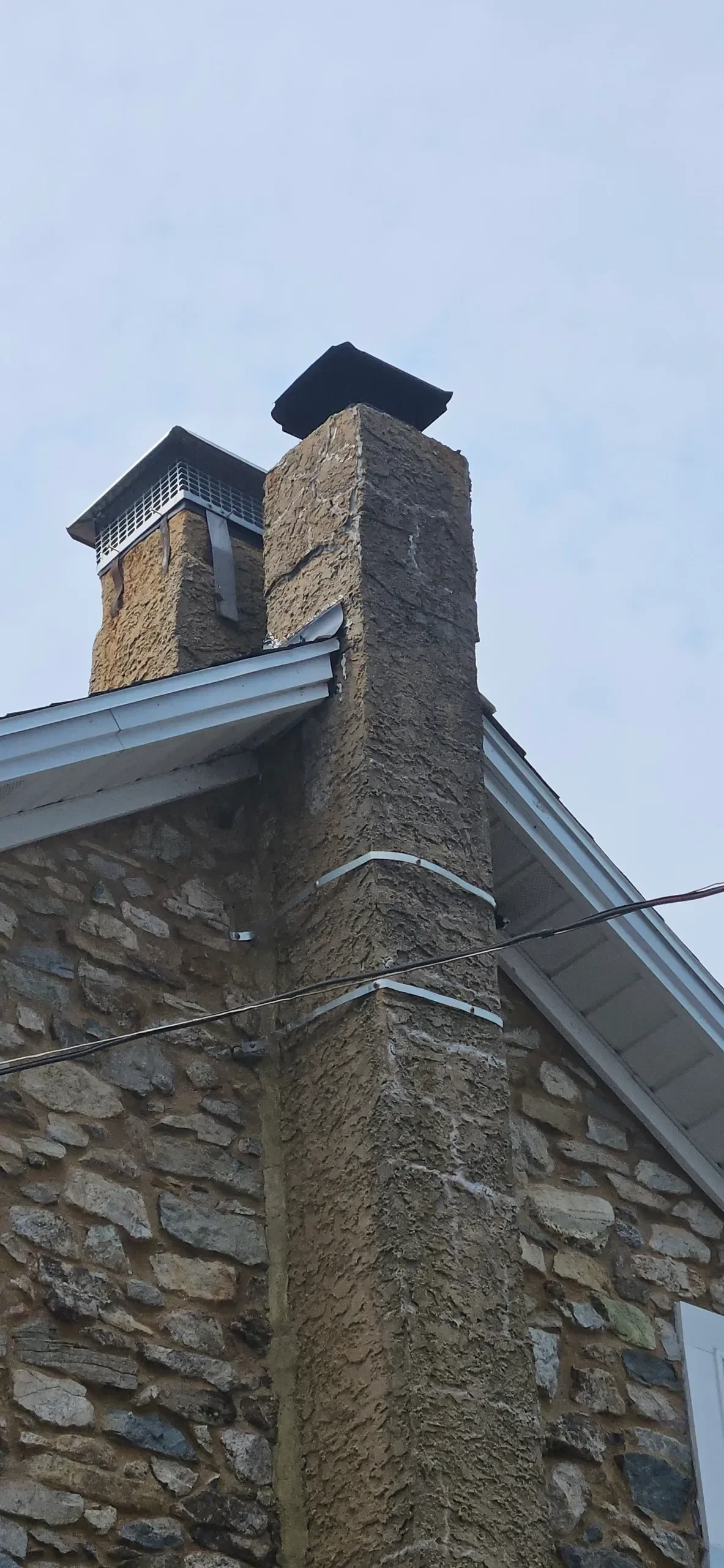 A low-angle view captures two textured stucco chimneys rising above a fieldstone wall and white roof eaves against a bright, overcast sky.
